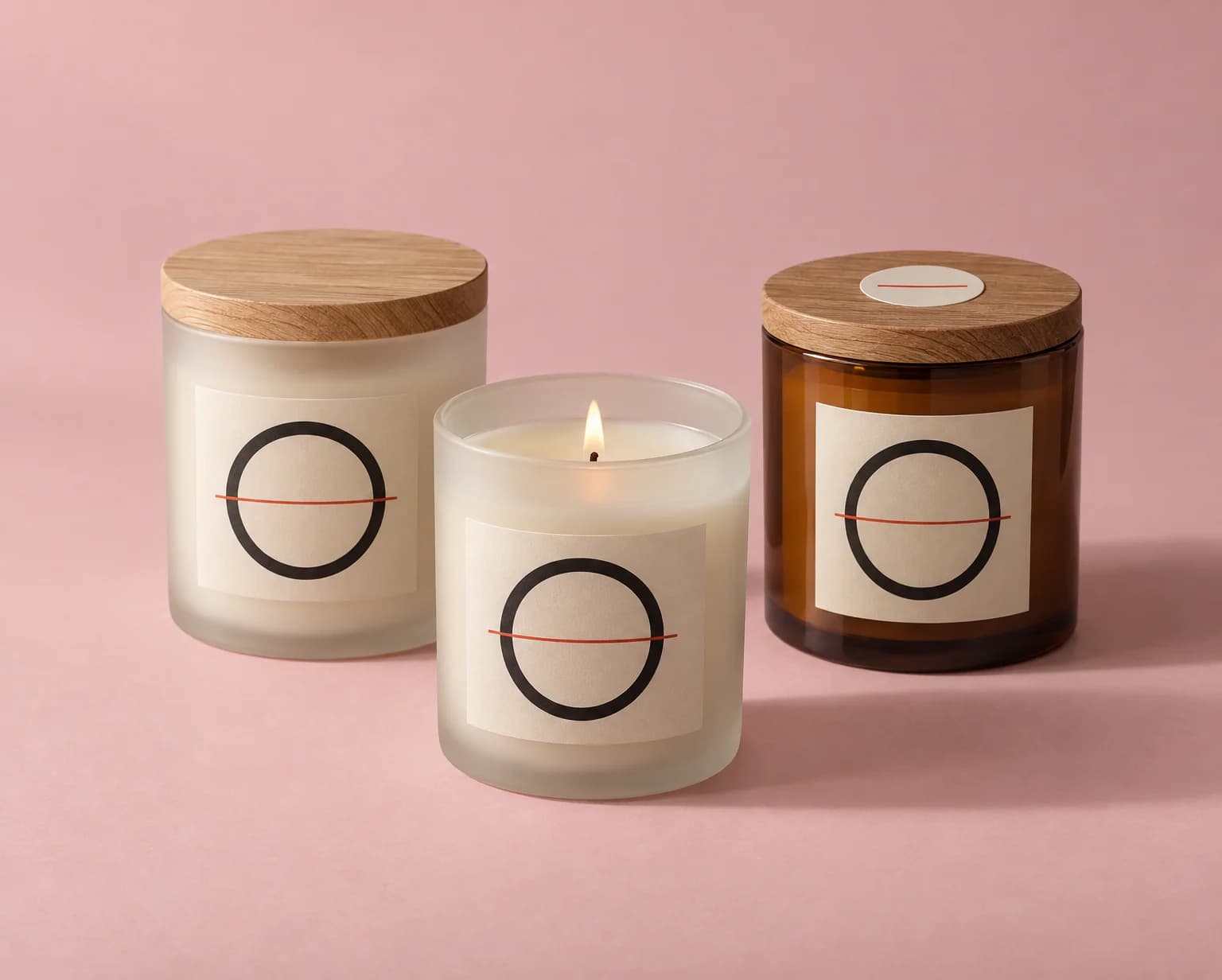 Two frosted glass candle jars and an amber candle jar with wooden lids, each with a matte cream label with a black ring, on a pink backdrop