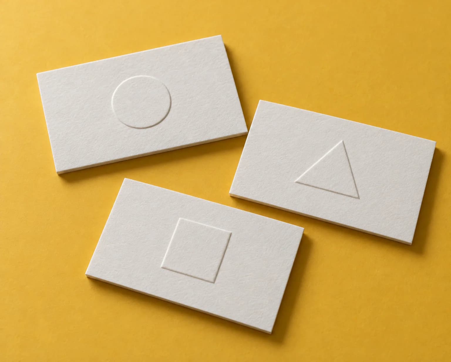 Three thick white business cards with embossed circle, triangle and square marks casting small shadows, on a mustard backdrop