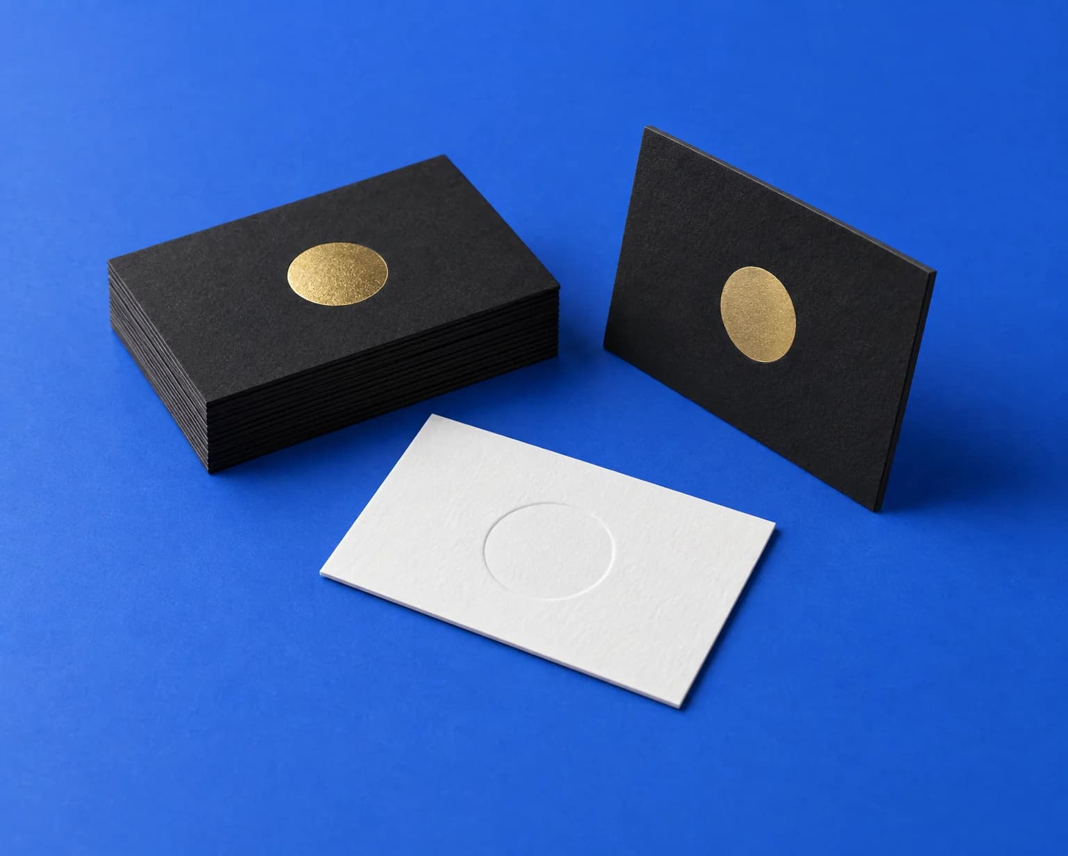 A stack of thick black business cards with a gold foil mark, one standing on its edge, and a white cotton card with an embossed circle, on a blue backdrop