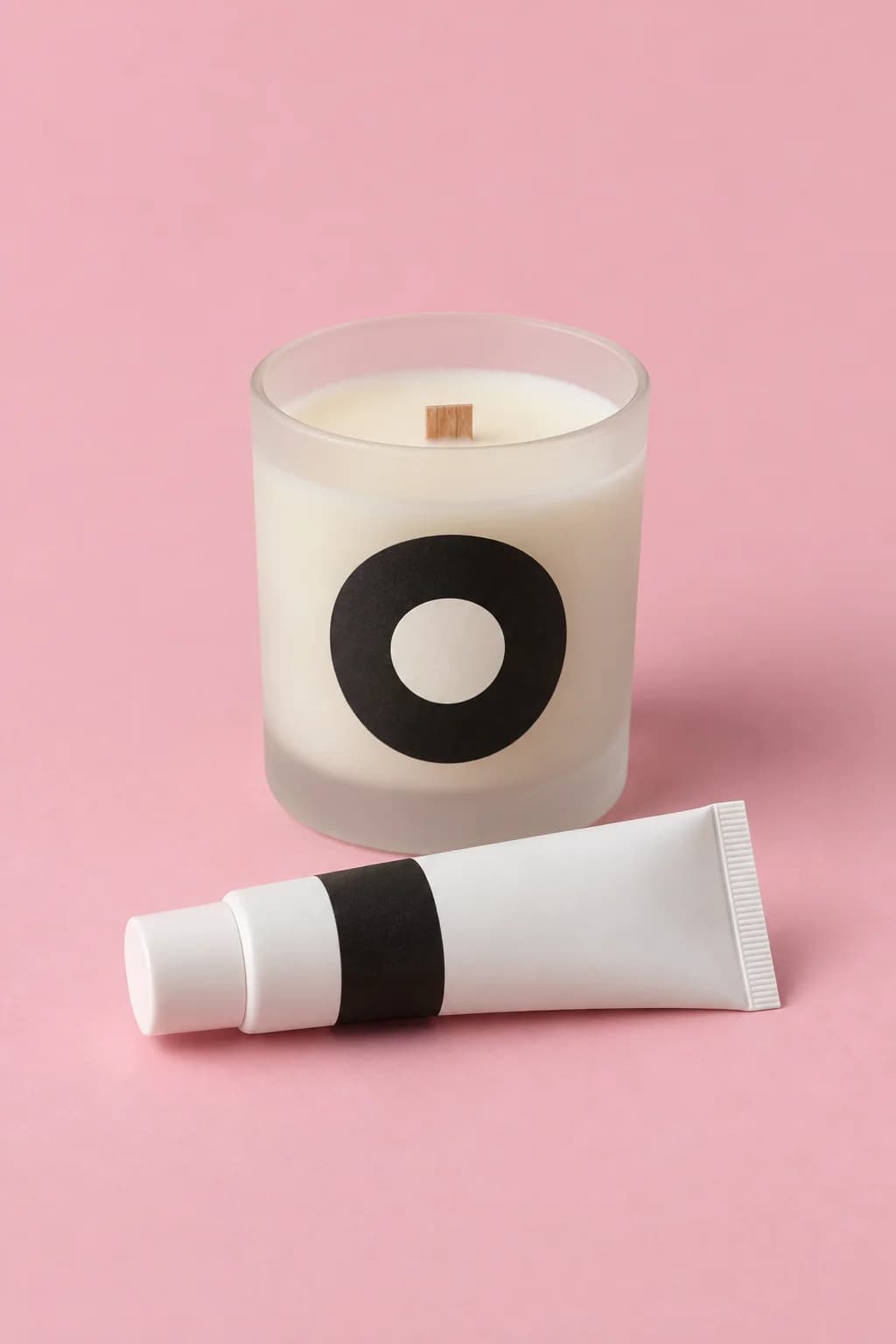 Frosted glass candle with a round black label beside a labelled white cosmetic tube on a blush pink backdrop